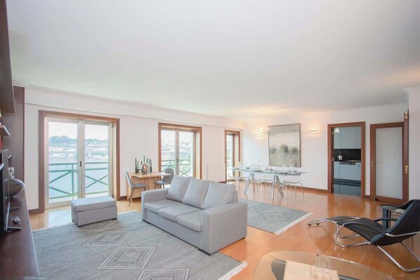 A modern living room with a sofa, a dining table, and a TV. There is a balcony with a view of the city.