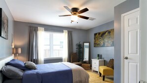 A bedroom with a bed, a ceiling fan, a window with curtains, a plant, a mirror, a dresser, and a door. - Serenity Haven with ShellCrestCorp Apt 106 (Memphis)