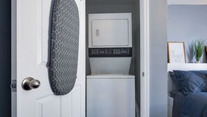 A compact laundry room with a washing machine and dryer, a patterned ironing board, and a bed with blue bedding in the adjacent room. - Serenity Haven with ShellCrestCorp Apt 106 (Memphis)
