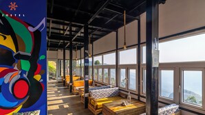 A hostel interior with wooden benches and tables, large windows offering views, and a colorful mural on the left. - Zostel Plus Poombarai (Kodaikanal) (Kodaikanal)
