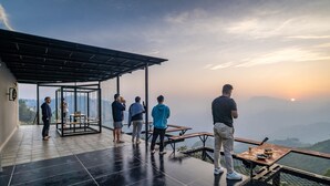 A terrace with a glass enclosure, a table with a drink, and a view of the mountains. - Zostel Plus Poombarai (Kodaikanal) (Kodaikanal)