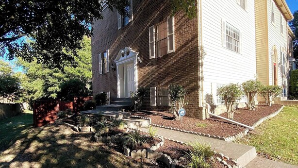 A two-story brick house with a white front door, a small front porch, and a well-maintained garden with a stone pathway and various plants. - Room in House - Majestic Private Room (Alexandria)