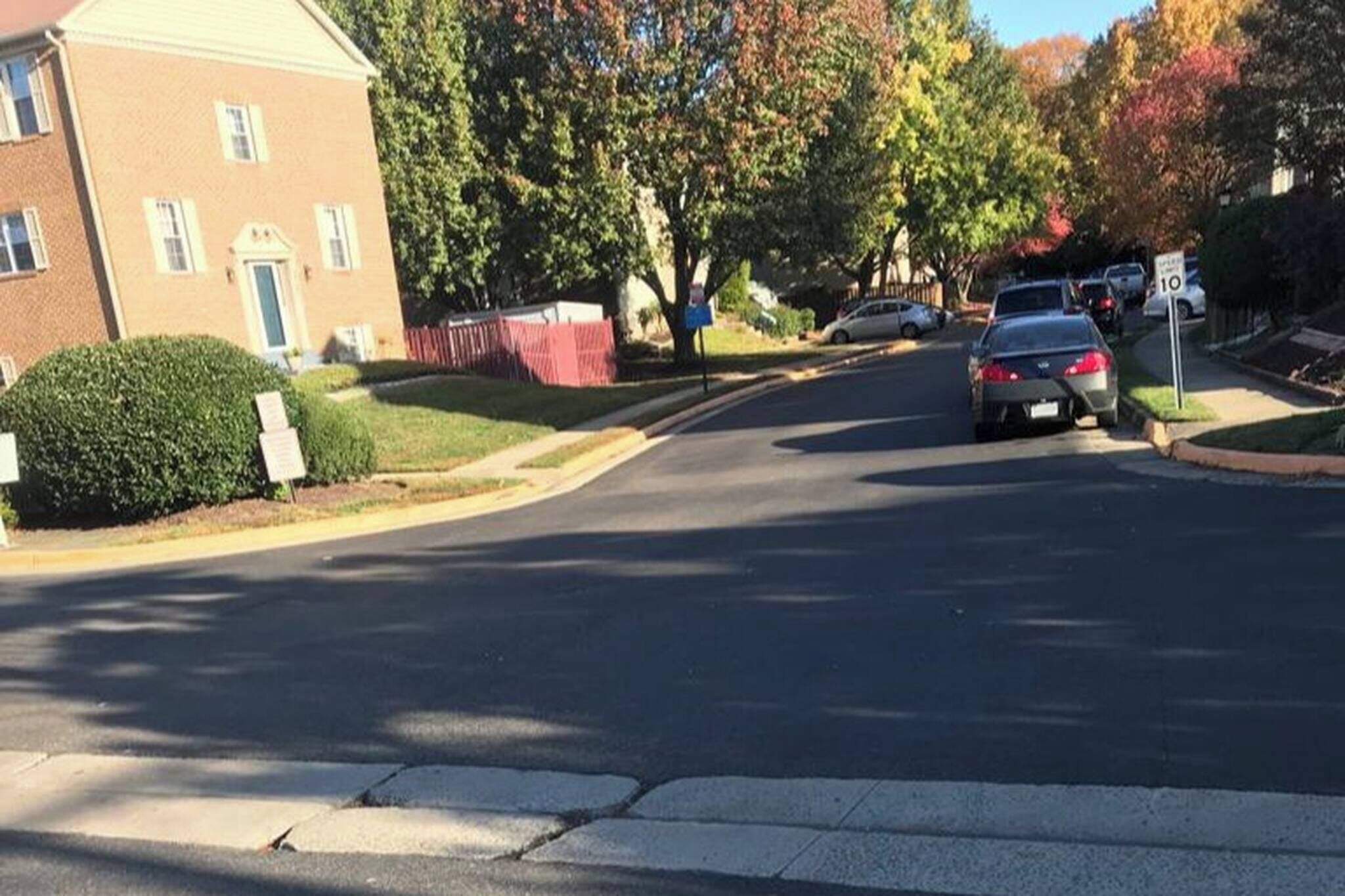 A residential street with a brick building on the left, parked cars, and trees with autumn foliage.