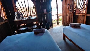 Una habitación con dos camas, cada una con una toalla que dice "Shambhala". Hay un escritorio de madera con un ventilador y una mesita. La habitación tiene una estructura de madera rústica con vistas al exterior.