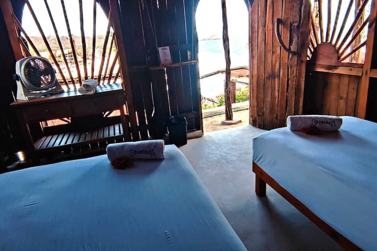 A room with two beds, each with a towel labeled "Shambhala." There is a wooden desk with a fan and a small table. The room has a rustic wooden structure with a view outside.