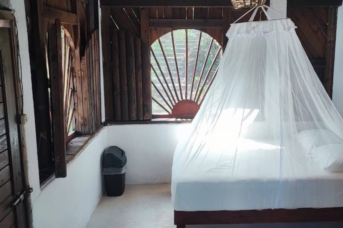 A room with a bed under a white canopy, wooden walls, a window with a view, and a small trash bin.