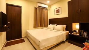 A hotel room with a bed, a wardrobe, a television, an air conditioning unit, and a nightstand with a lamp and books. - Blu Ivy Serene Serviced Apartments (Chennai)