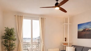 A modern living room with a grey sofa, two white chairs, a small round table, a ceiling fan, a large window with curtains, a potted plant, and a painting on the wall. - Modern flat near Bercy (Paris)