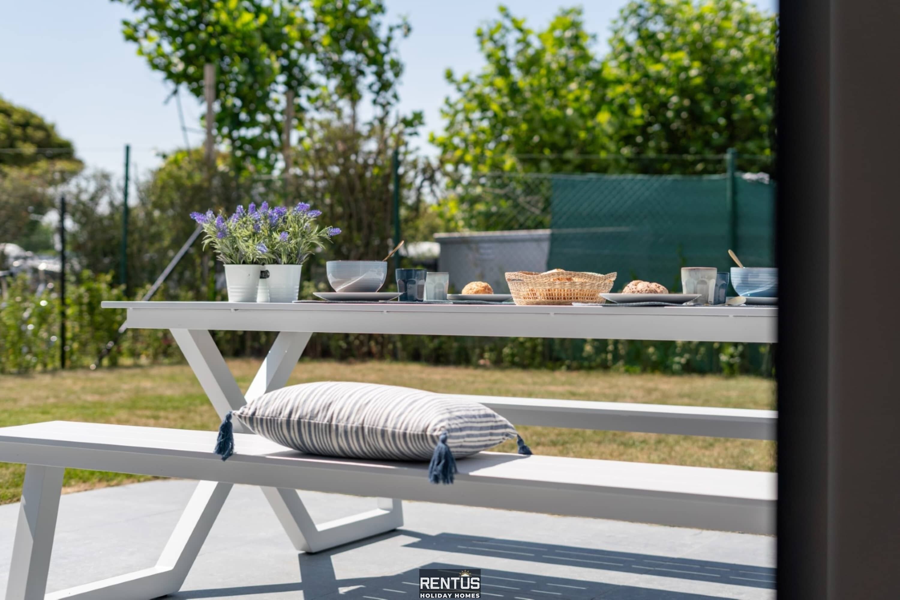 Een witte picknicktafel met een bankje, een bloempot en een mand.