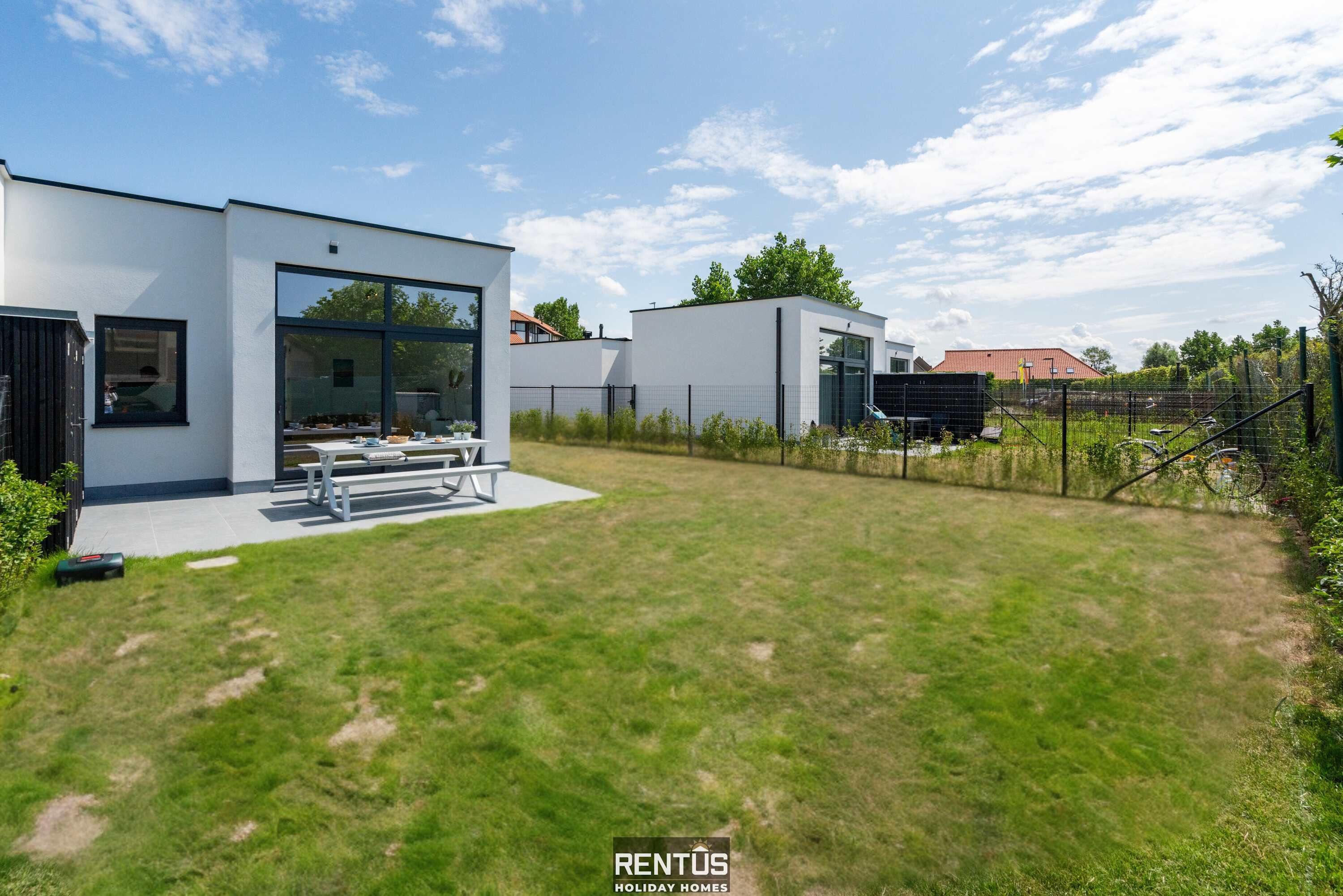 A modern two-story house with a large glass door entrance, a patio with outdoor furniture, and a fenced backyard.