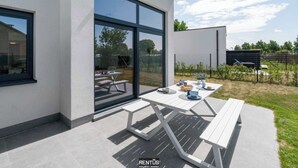 Uma área de pátio moderna com mesa e bancos brancos, amplas portas de vidro e vista para a paisagem ao redor.