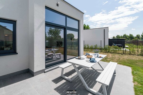 A modern patio area with a white table and benches, large glass doors, and a view of the surrounding landscape.