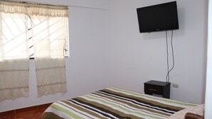 Une chambre à coucher avec un lit, un téléviseur fixé au mur, une petite table de chevet et une fenêtre avec des rideaux.