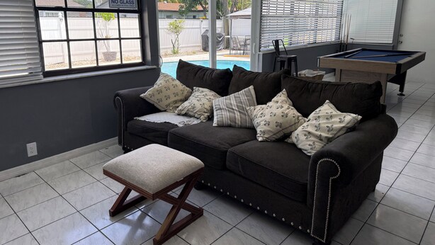 A room with a dark-colored sofa, a wooden bench, a pool table, and a view of a backyard with a pool.