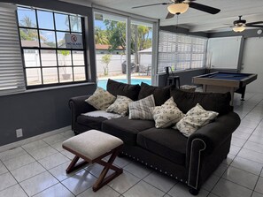 A room with a dark-colored sofa, a wooden bench, a pool table, and a view of a backyard with a pool.