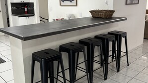 A kitchen with a central island, black bar stools, a fire extinguisher, a coffee maker, and a framed artwork on the wall.