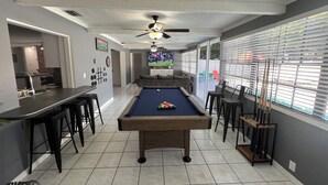 A pool table in a room with a ceiling fan, a bar area, and a view through large windows.