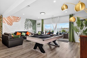 A modern living room with a pool table, a sofa with cushions, and a large window offering a view. - Luxury Desert Hot Springs 4BR/3BA Vacation Home /Private Pool & Mountain Views (Desert Hot Springs)