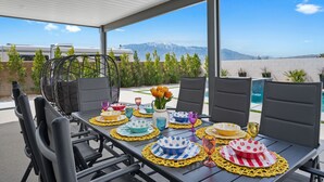 Un área de comedor al aire libre con vistas a montañas, equipada con sillas negras y una mesa dispuesta con platos y cristalería de colores.