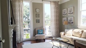 A living room with a sofa, armchair, and coffee table, a patterned curtain, and a view of the outdoors. - Garden District | Steps to St. Charles Streetcar (New Orleans)