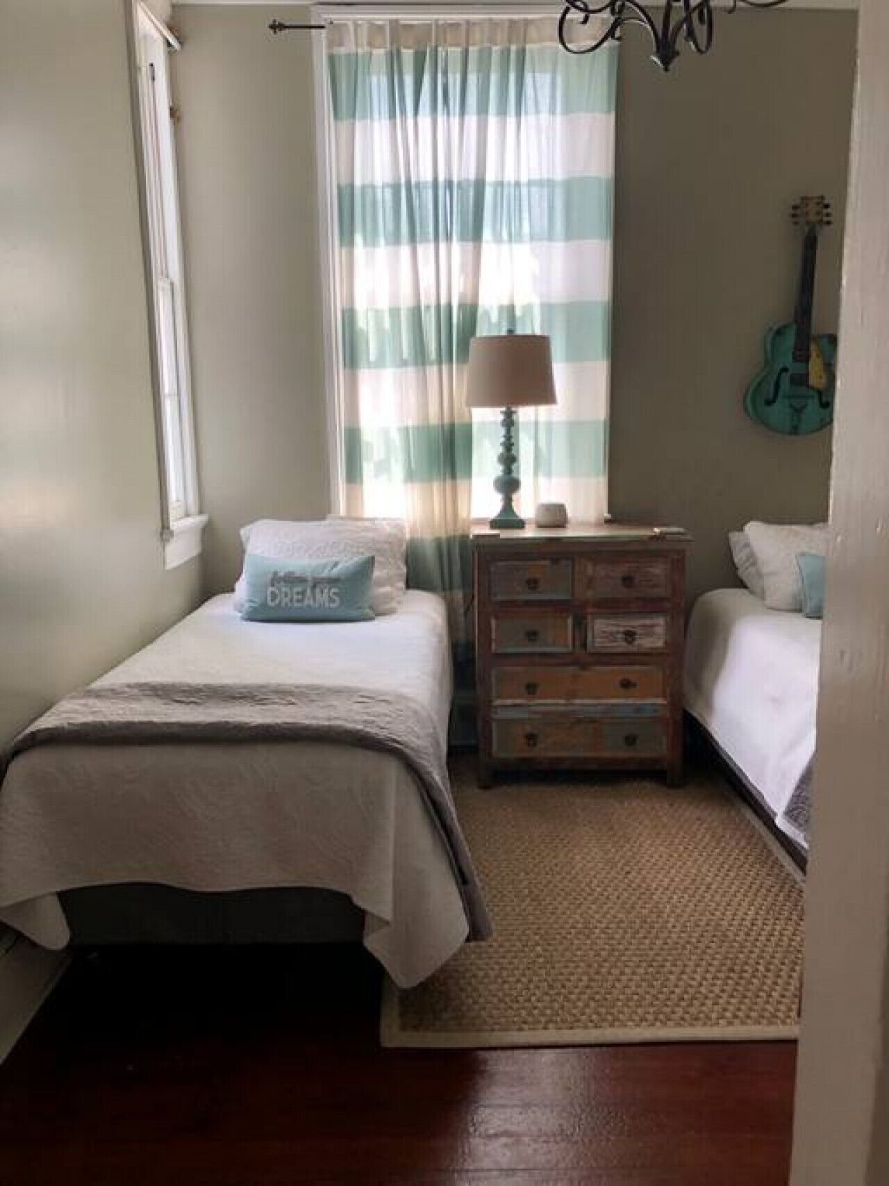 Une chambre avec deux lits, une table de chevet avec des tiroirs, une lampe, une guitare et une fenêtre avec des rideaux.