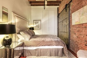 A bedroom with a bed, a nightstand, a lamp, a framed picture on the wall, and a brick wall.