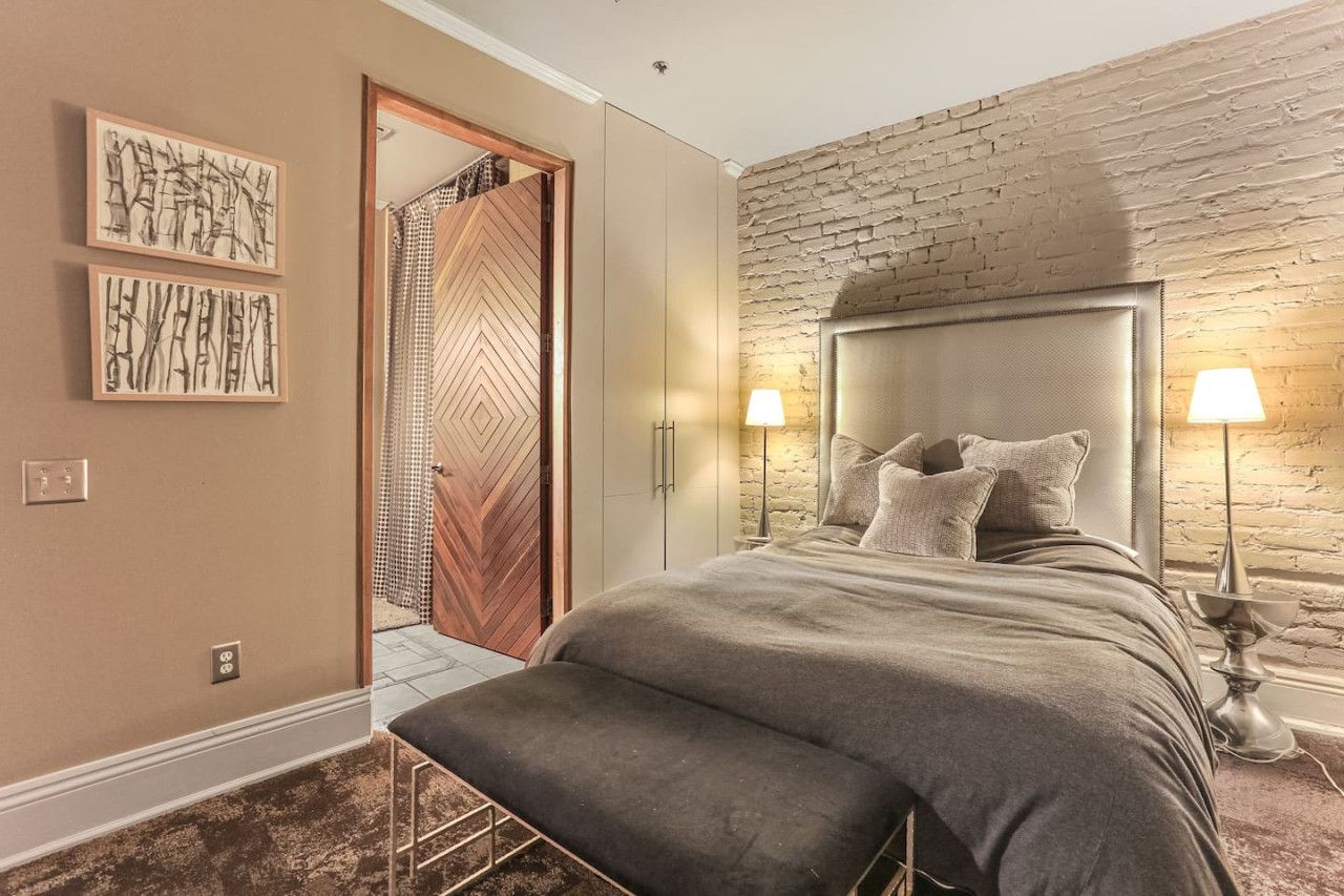 Un dormitorio con una cama grande, dos lámparas de noche, un banco, una puerta de madera y una pared de ladrillo.