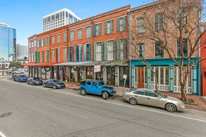A street view with a row of buildings, including a red brick building with green shutters and a blue SUV parked in front.