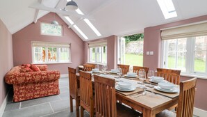 A dining area with a wooden table set for six, surrounded by wooden chairs, with a red patterned sofa against the wall and a window overlooking a garden. - The Old Bakehouse (Marlborough)