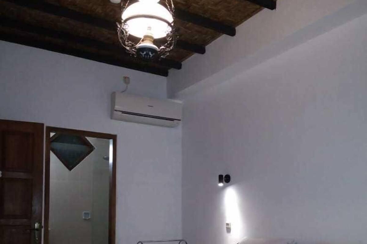 A bedroom with a wooden bed frame, patterned bedding, a hanging light fixture, a door, and an air conditioning unit.