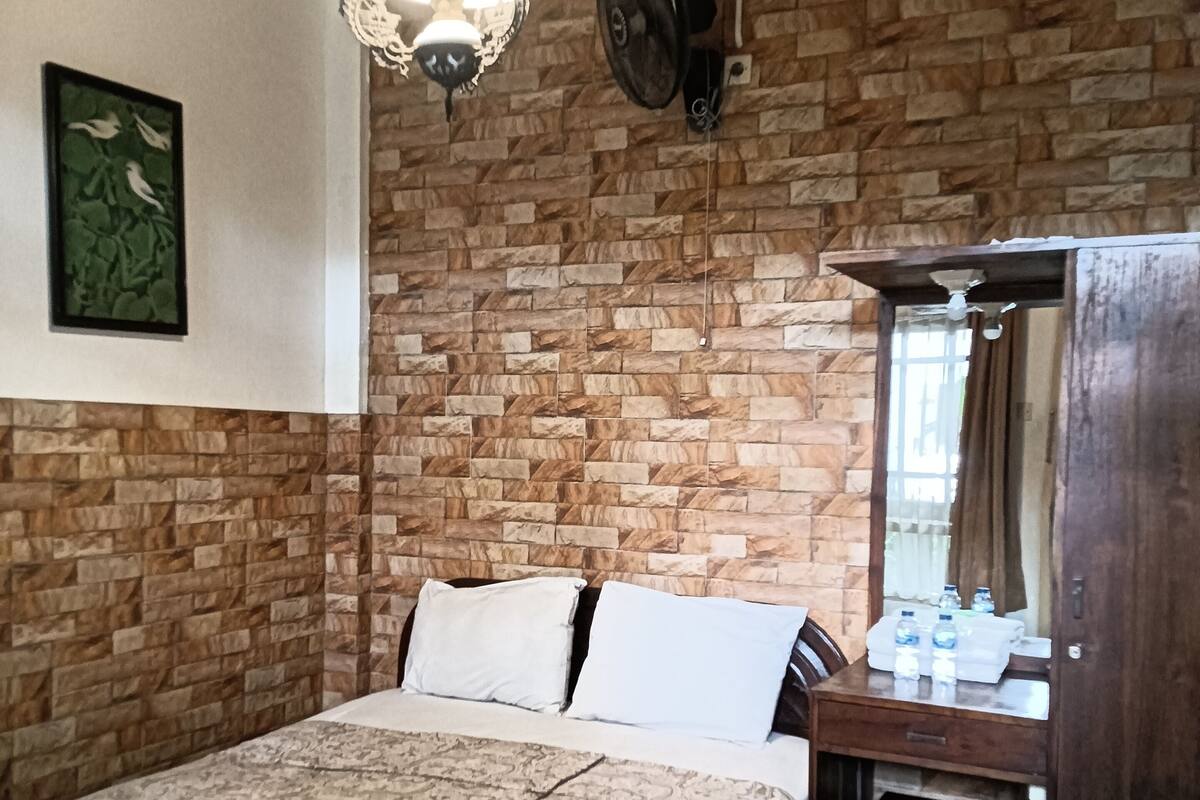 A bedroom with a bed, a desk, a chair, a hanging light fixture, and a brick wall.