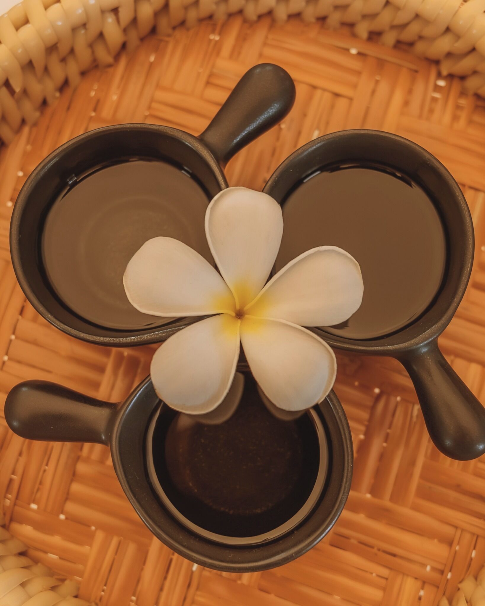 En bild tagen ovanifrån som visar en flätad korg med tre svarta muggar med handtag. Varje mugg är fylld med en mörk vätska, och i mitten av korgen står en vit blomma med ett gult mittparti.