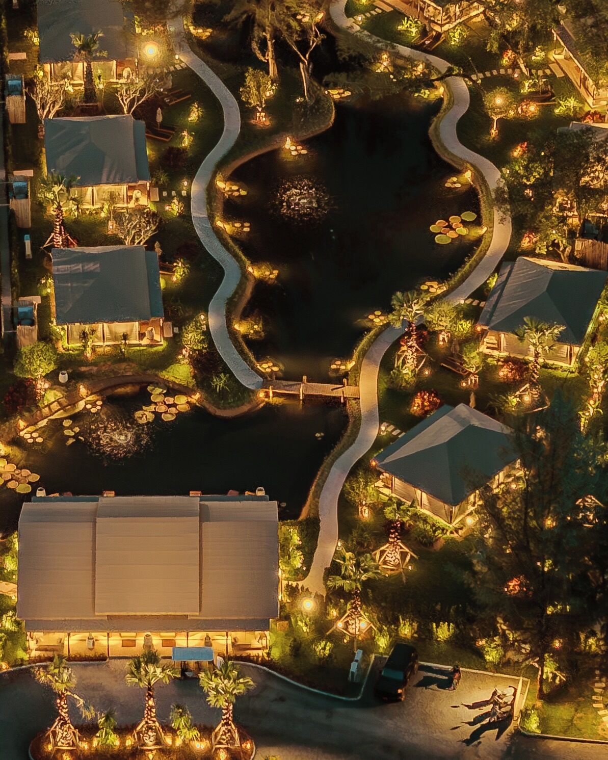 An aerial view of a resort with a central water feature, connected by winding pathways, surrounded by well-lit buildings and lush landscaping.