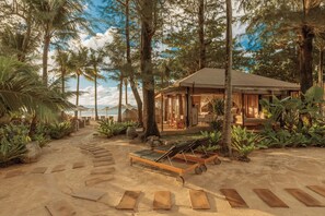 Un resort frente a la playa con una terraza de madera que da a un espacio de descanso con sillones, rodeado de exuberante vegetación tropical y un gran árbol.