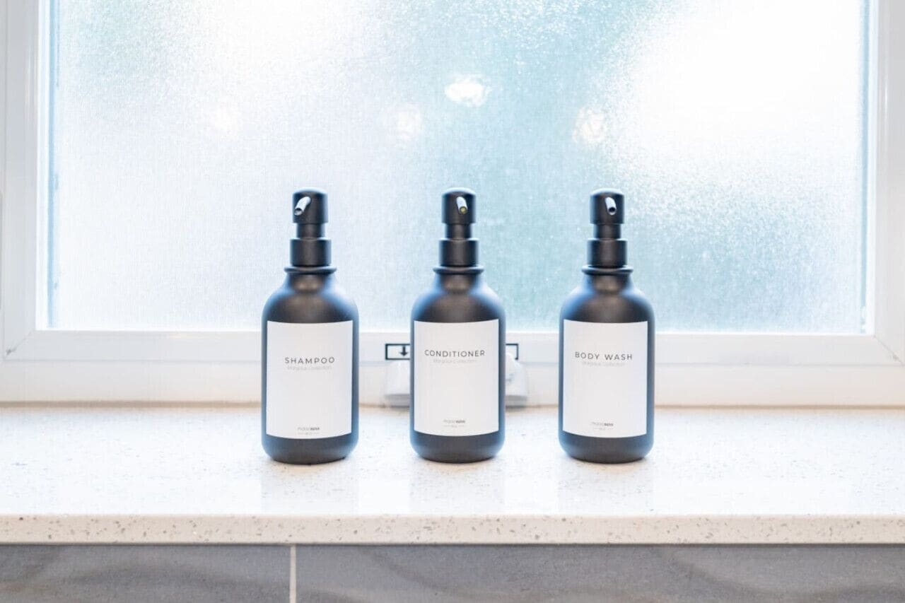 Three bottles of shampoo, conditioner, and body wash on a countertop by a window.