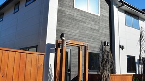 Una casa moderna de dos pisos con fachada gris y blanca, cerca con valla de madera y un cielo azul despejado.