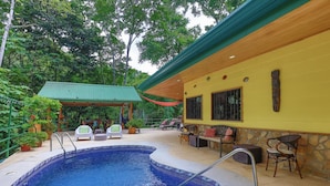Una piscina con bordo piastrellato, circondata da zone relax all'aperto e un'area coperta con tetto verde.