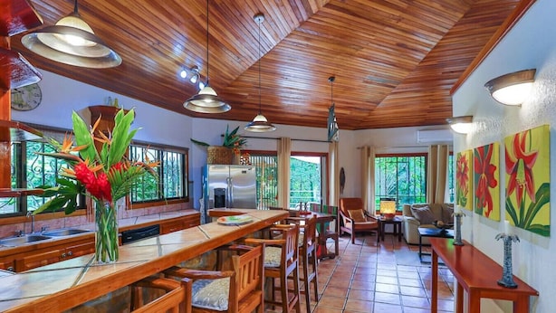 Cucina spaziosa con mobili in legno, un lungo tavolo da pranzo e sedie. Sono presenti luci a sospensione e un'ampia finestra con vista su un'area verde.