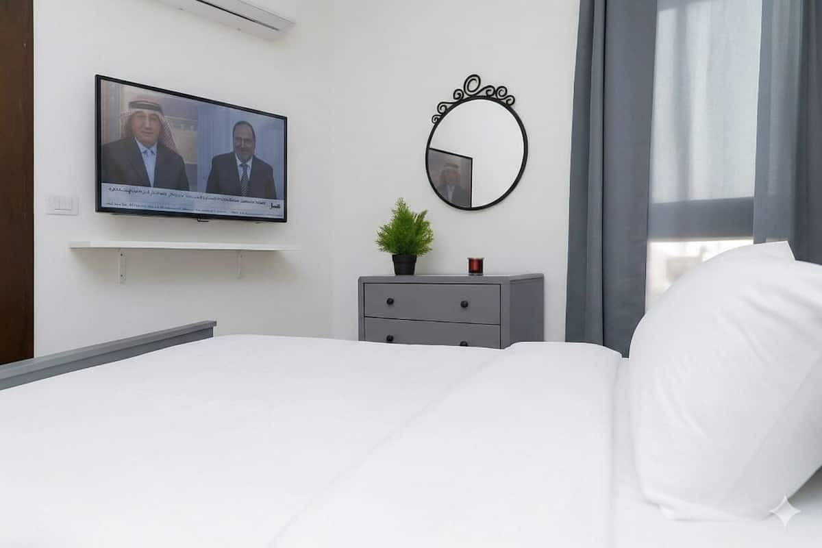 A hotel room with a bed, a television mounted on the wall, a small plant on a dresser, and a round mirror.