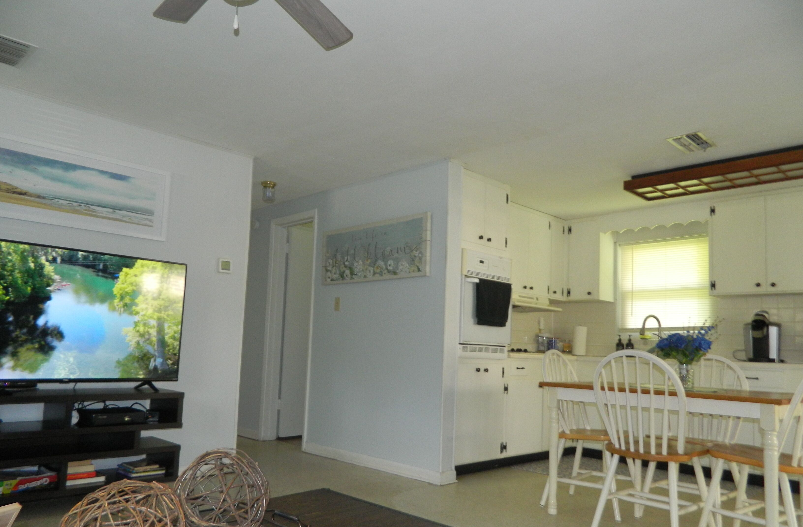 A modern kitchen with white cabinets, a dining table with chairs, a television, and a ceiling fan.