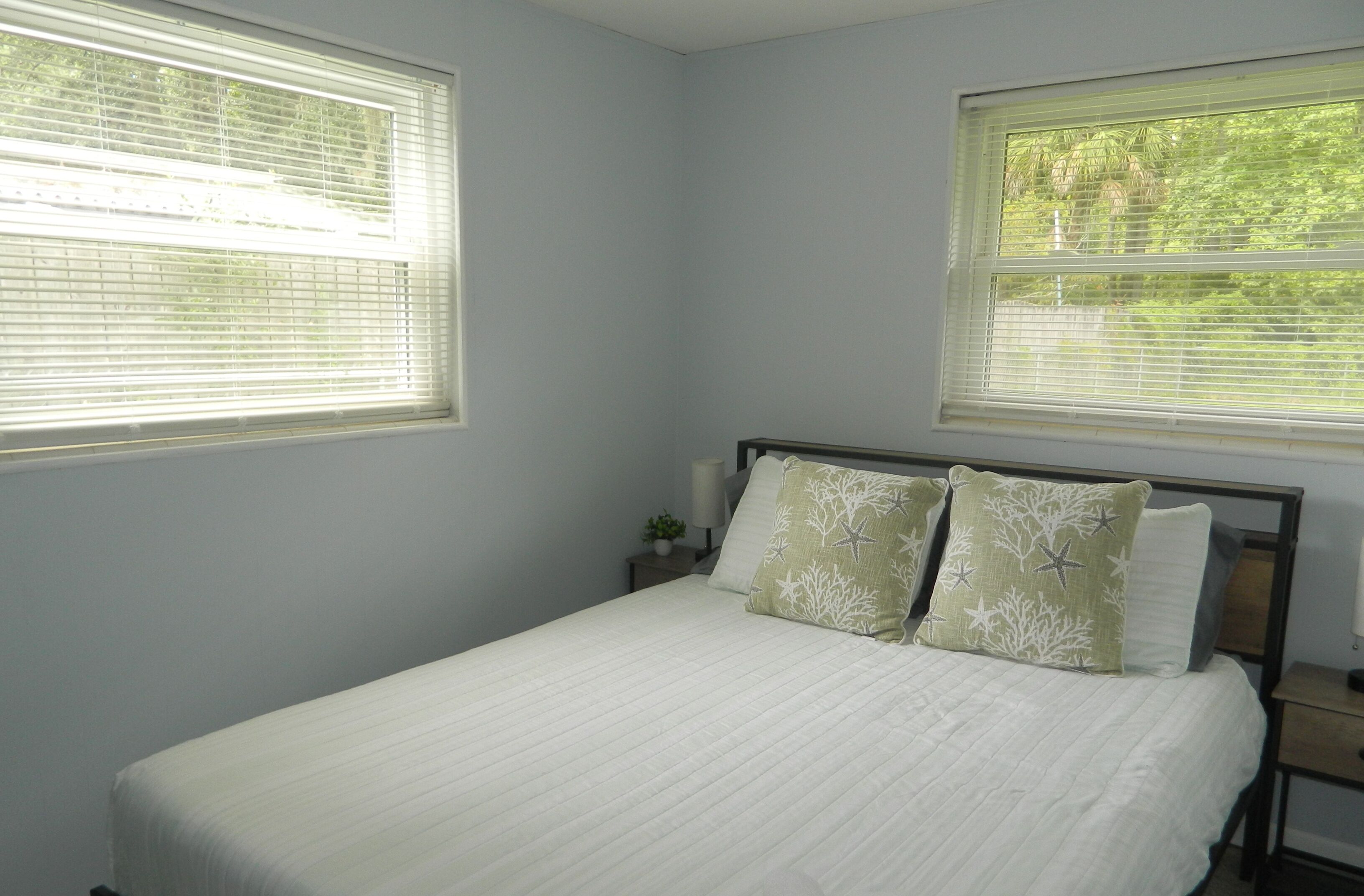 A bedroom with a bed, two nightstands, a lamp, and two windows with blinds.