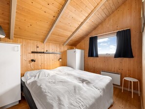 Quarto com painéis de madeira, uma cama, um frigorífico branco pequeno, uma estante com uma placa e uma janela com cortinas, além de um banquinho.