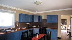 Cozinha com armários azuis, uma mesa de jantar com cadeiras, uma janela com cortinas e vista para outro cômodo.