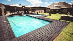 Piscina com deck de madeira, construções com telhado de palha e céu azul e límpido.