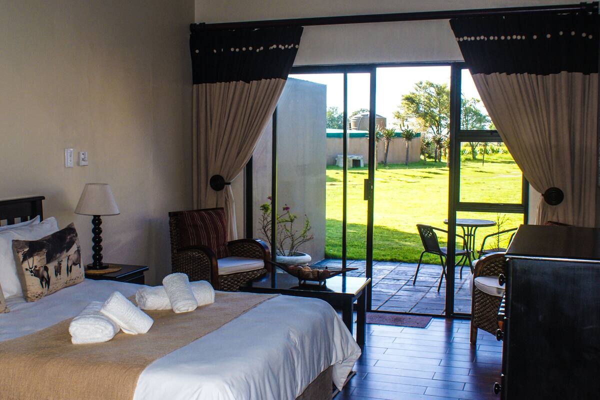 A hotel room with a bed, a nightstand, a lamp, a chair, and a view of a grassy yard through a glass door.