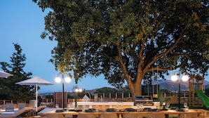 An outdoor dining area with a large table set for six, surrounded by wicker chairs, under a tree. - Heated pool, outdoor playground, convenient location close to the beach and town (Rethymno)