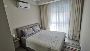 Ein Hotelzimmer mit Bett, Nachttisch, Klimaanlage, einem Fenster mit leichtem Vorhang und Blick ins Freie.