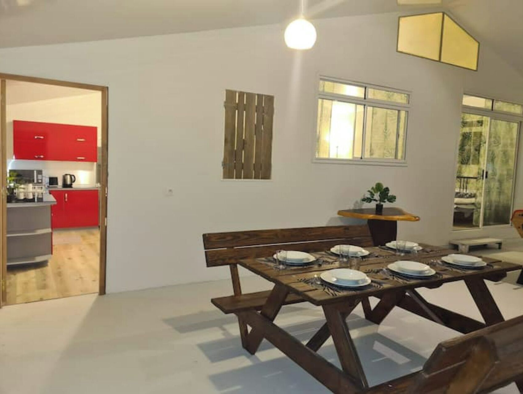A dining area with a wooden table set for four, a bench, and a small plant on a side table. A kitchen with red cabinets is visible in the background.