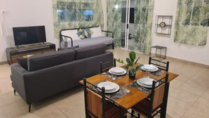 A modern living room with a sofa, a TV, a dining table set for four, and a bed with curtains. - "Papara - Taharuʻu Beach 2BR House - Surf & Family - garden & secure garage (Papara)