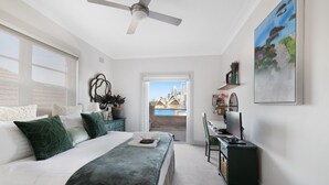 Ein Schlafzimmer mit einem Bett, einem Schreibtisch, einem Stuhl, einem Deckenventilator, einem Gemälde und Blick auf die Stadt durch ein Fenster.